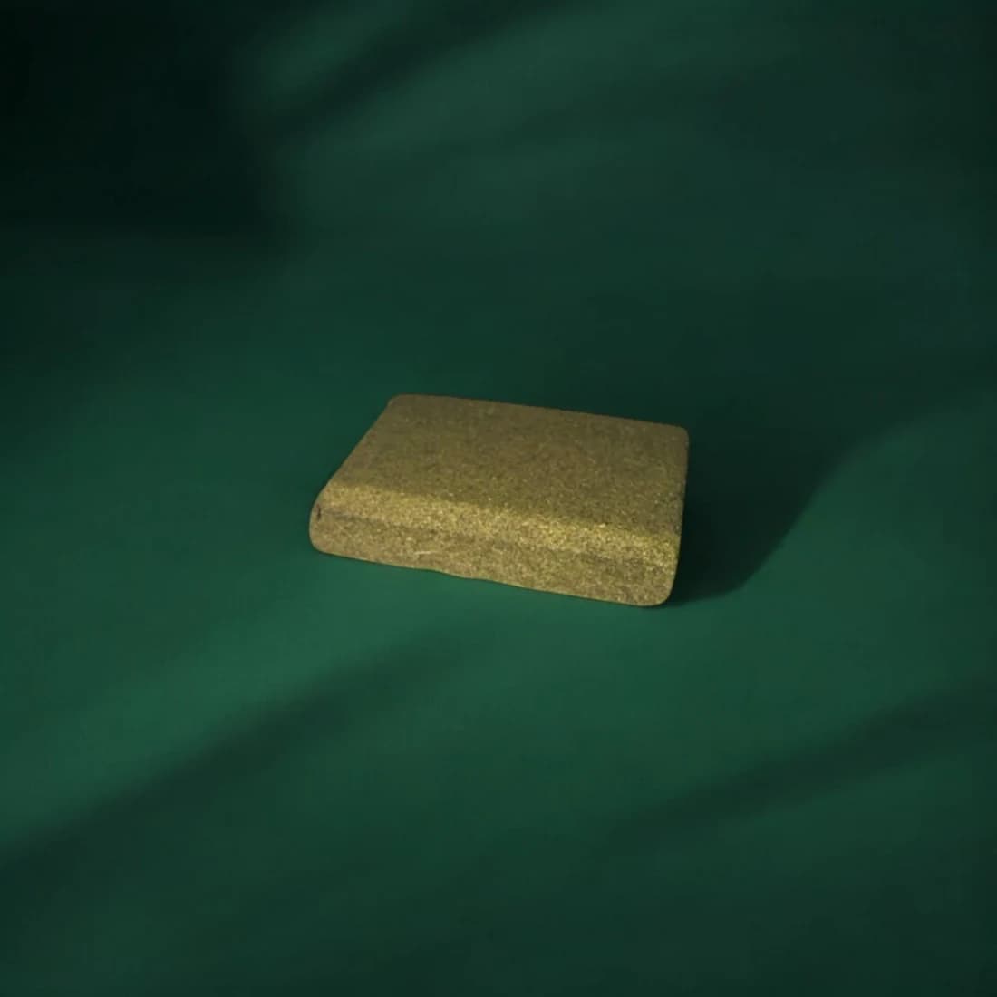 A bare Professor Herb hemp briquette out of its pot on a dark green cloth, showing the dense granular pressed texture and rounded corners